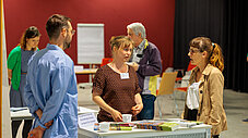 Drei Personen im Gespräch an einem Ausstellungstisch mit Büchern und einem Schild des Landesamts für Umwelt, weitere Personen im Hintergrund.