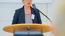 Frau mit Brille spricht an Rednerpult vor einer Präsentationsleinwand