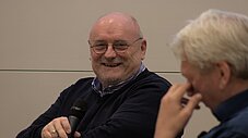 Person hält Mikrofon und lacht in einem Gesprächsformat, eine weitere Person ist im Vordergrund unscharf sichtbar