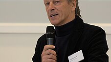 Person spricht mit Mikrofon in der Hand an einem Stehtisch vor einer Leinwand
