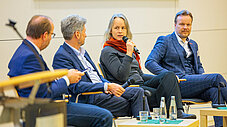 Vier Personen sitzen auf einem Podium, eine spricht ins Mikrofon bei einer Veranstaltungsdiskussion.