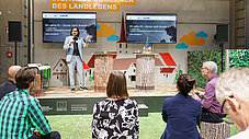 Vortragender auf Bühne bei Smart Country Brandenburg, Bildschirme zeigen Folie zu 60 Jahre KI, Publikum davor