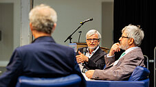 Drei Personen in Sesseln im Gespräch auf einer Podiumsdiskussion, eine Person spricht ins Mikrofon