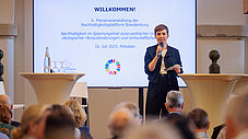 Moderatorin spricht mit Mikrofon vor Leinwand zur 6. Plenarveranstaltung der Nachhaltigkeitsplattform Brandenburg