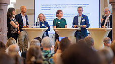 Sechs Personen auf dem Podium vor Leinwand mit Veranstaltungstext, Publikum im Vordergrund