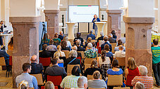 Publikum sitzt in einem historischen Saal und hört einer Rednerin am Podium zu