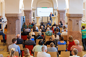 Publikum sitzt in einem historischen Saal und hört einer Rednerin am Podium zu