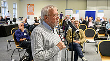 Person steht an einem Standmikrofon und spricht vor sitzendem Publikum in einem Konferenzsaal.
