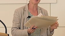 Person steht mit Unterlagen in der Hand und lächelt, Ansteckmikrofon sichtbar
