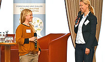 Zwei Frauen stehen mit Mikrofonen auf einer Bühne vor einem Nachhaltigkeitsbanner