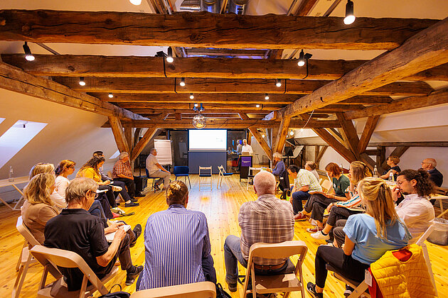 Gruppe sitzt in einem Dachgeschossraum mit Holzgebälk und verfolgt eine Präsentation