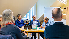 Gruppe diskutiert an einem Tisch mit Unterlagen, im Hintergrund ein Whiteboard in einem Gruppenarbeitsraum.